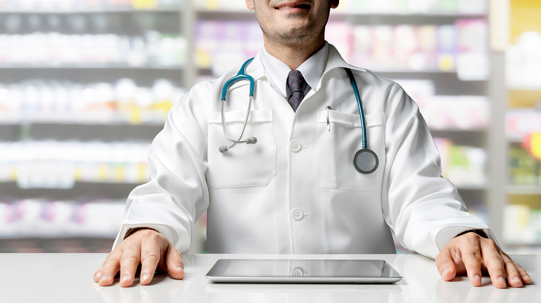 Male pharmacist sitting at table with tablet computer in pharmacy office. Medical healthcare staff and drugstore business.